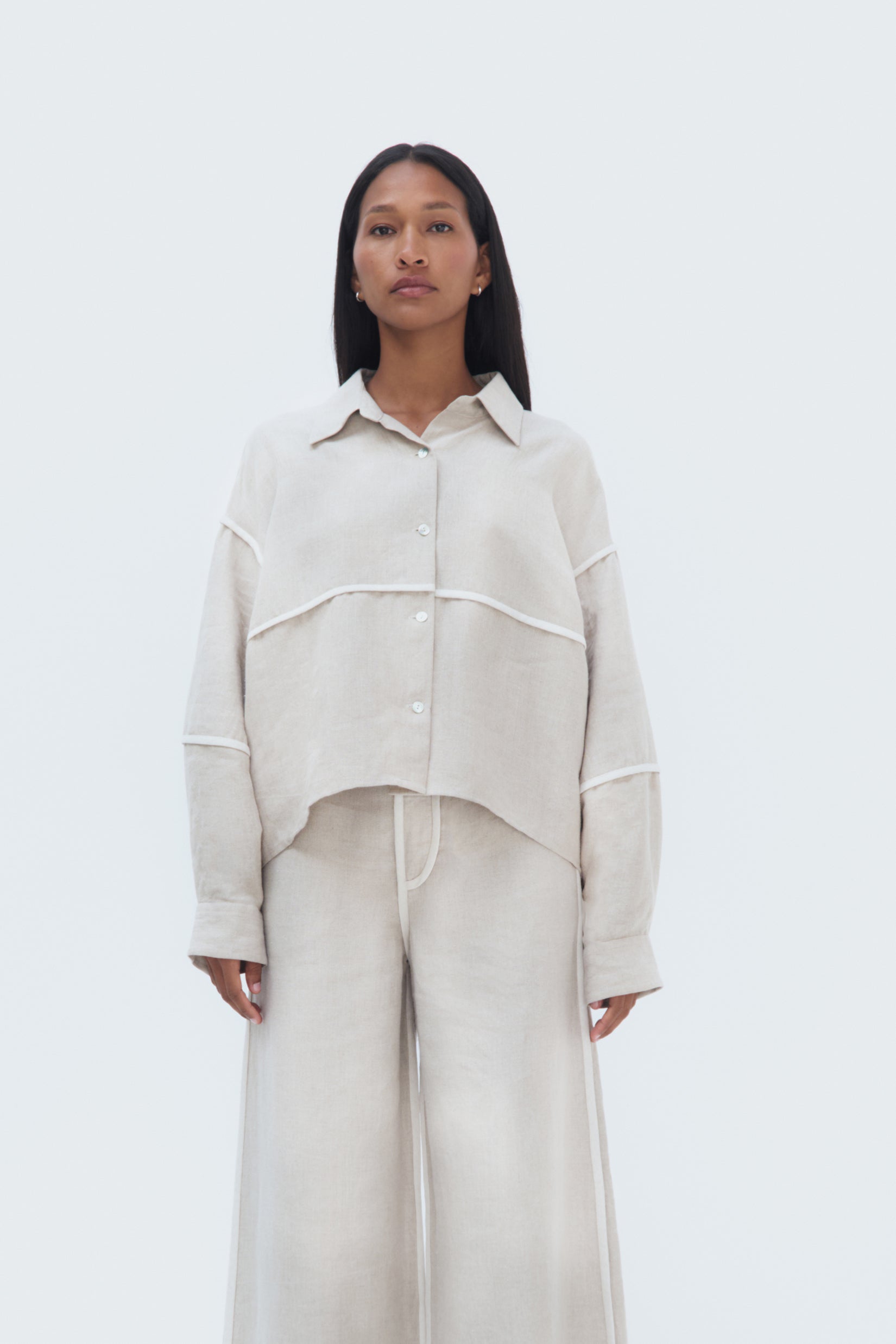 A woman modeling a beige linen blouse with piping details, paired with matching pants.