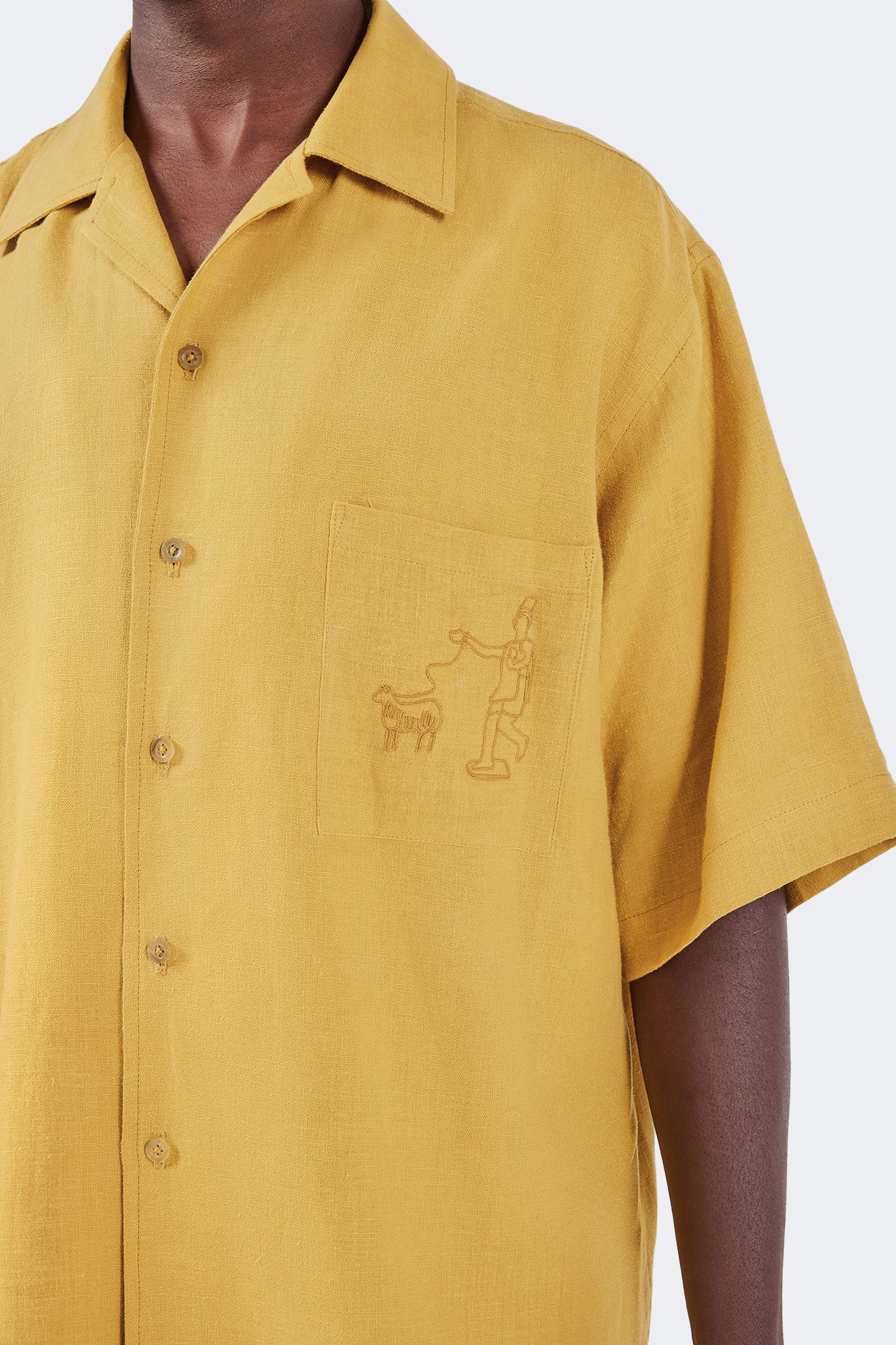 Angled detail of the Floreana Short-Sleeved Shirt in Mustard featuring the embroidered chest pocket, clean tailoring, and lightweight linen fabric.