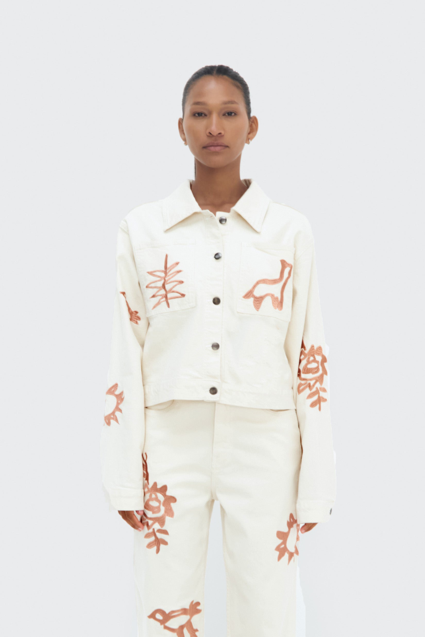 A model wearing a white cropped denim jacket with brown embroidery, paired with matching white pants.