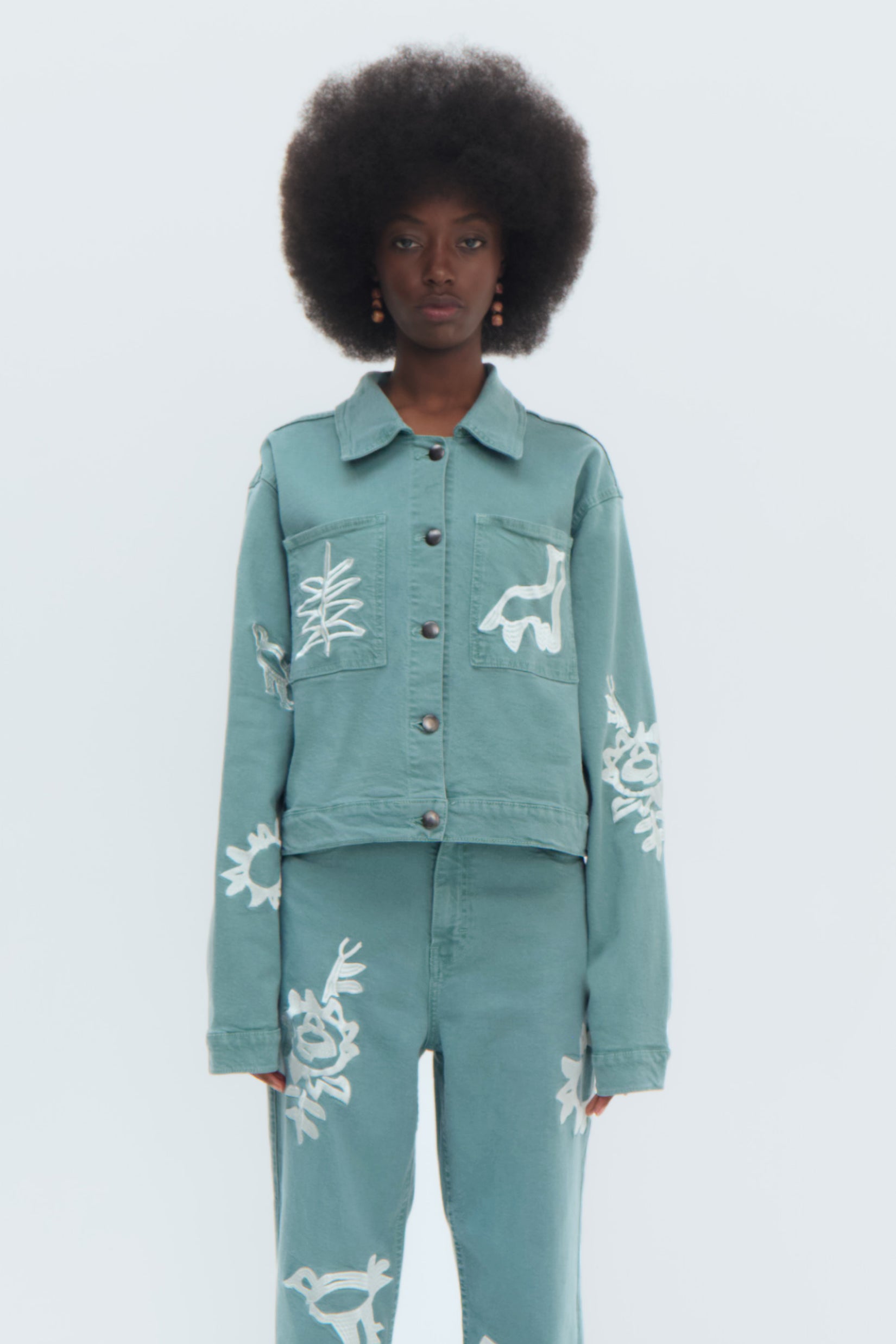 A woman modeling a green denim cropped jacket with white embroidery, featuring a fitted silhouette and button-up front.