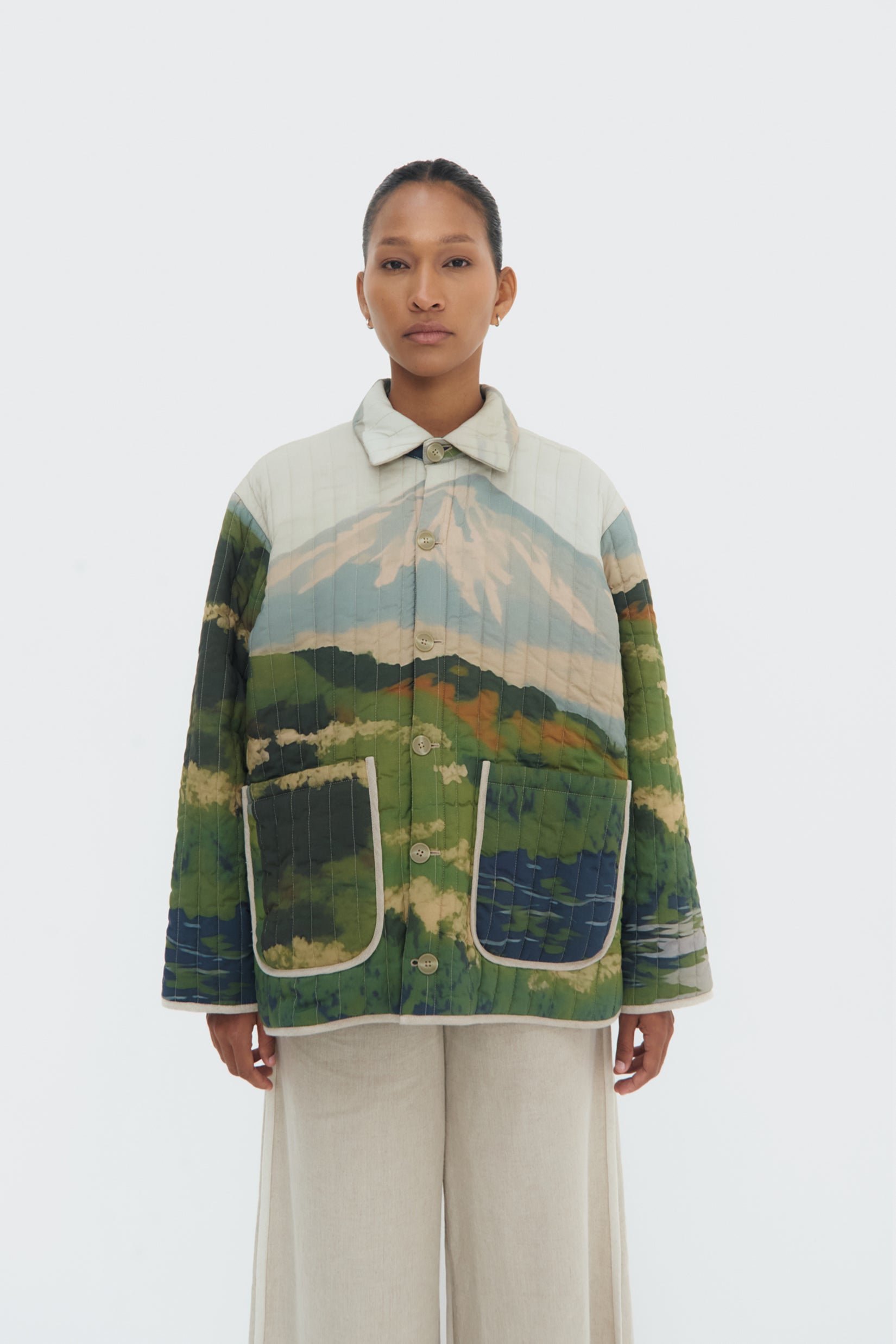A model wearing an oversized padded jacket with vertical stitching and an abstract print inspired by Andean landscapes. The jacket has a spread collar, button front closure, long sleeves, and two front patch pockets.