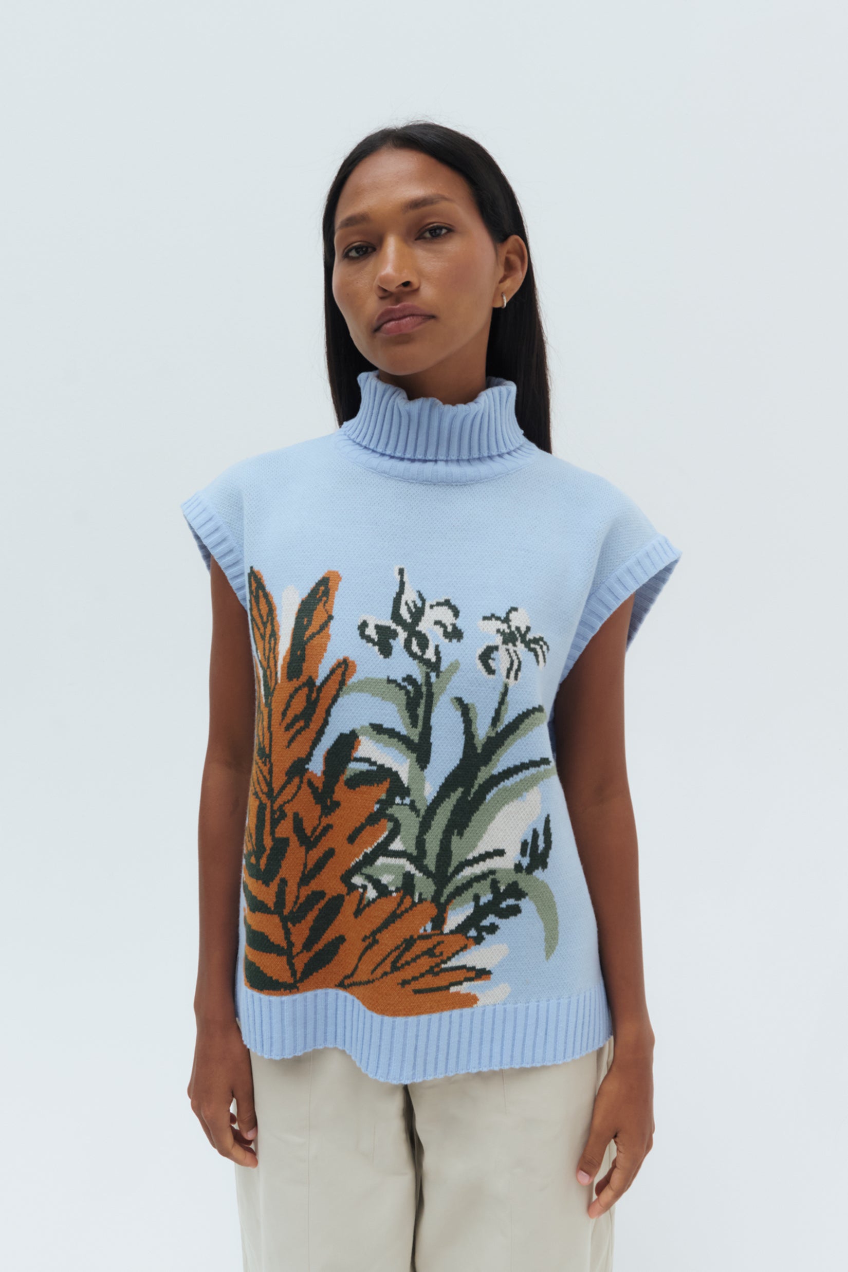 A woman wearing a sleeveless blue knit sweater vest with a floral design, paired with beige pants.