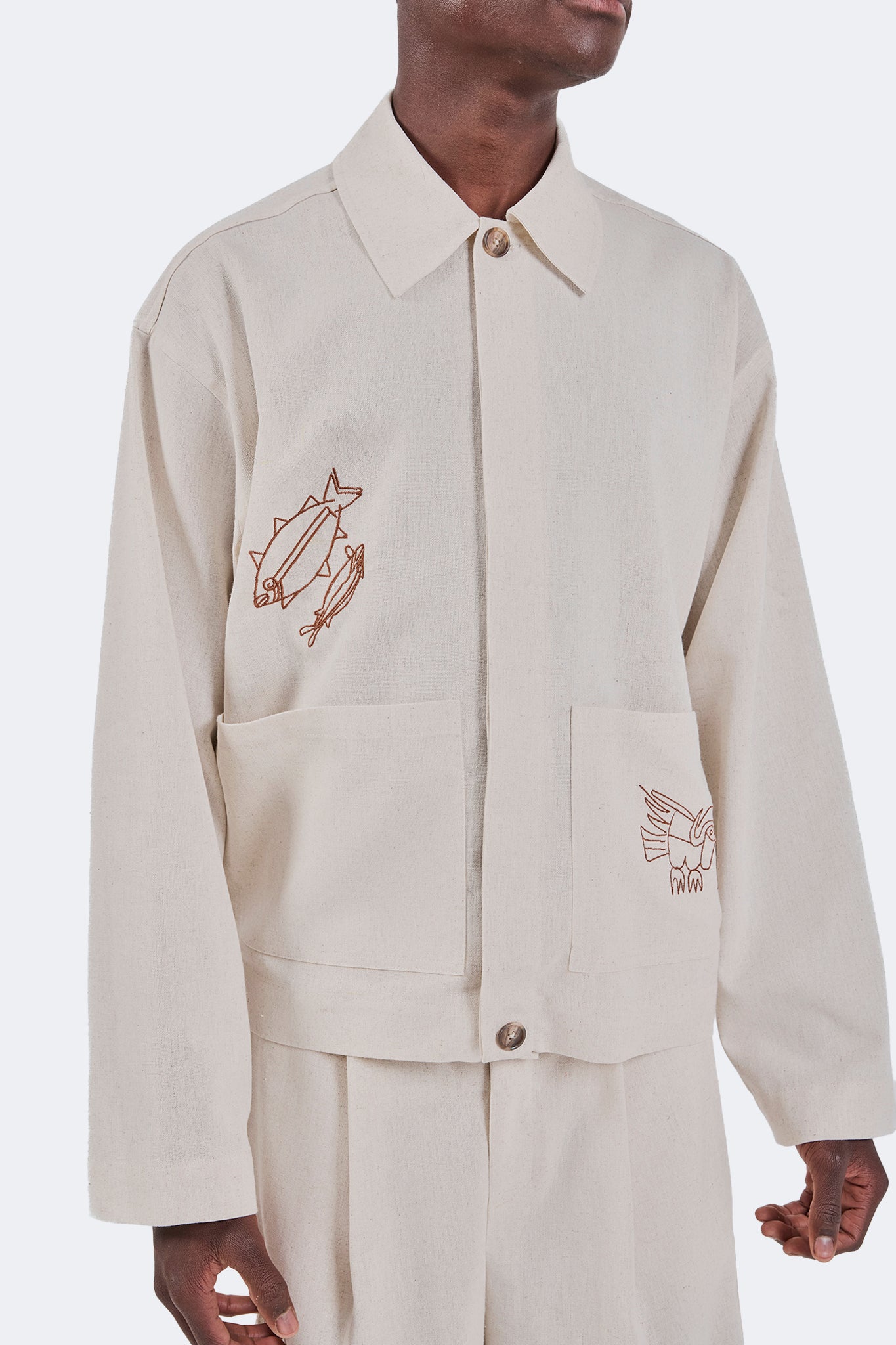Close-up of the Salango Linen Utility Jacket in Off-White highlighting the embroidered fish motif, oversized patch pockets, point collar, and natural linen texture.