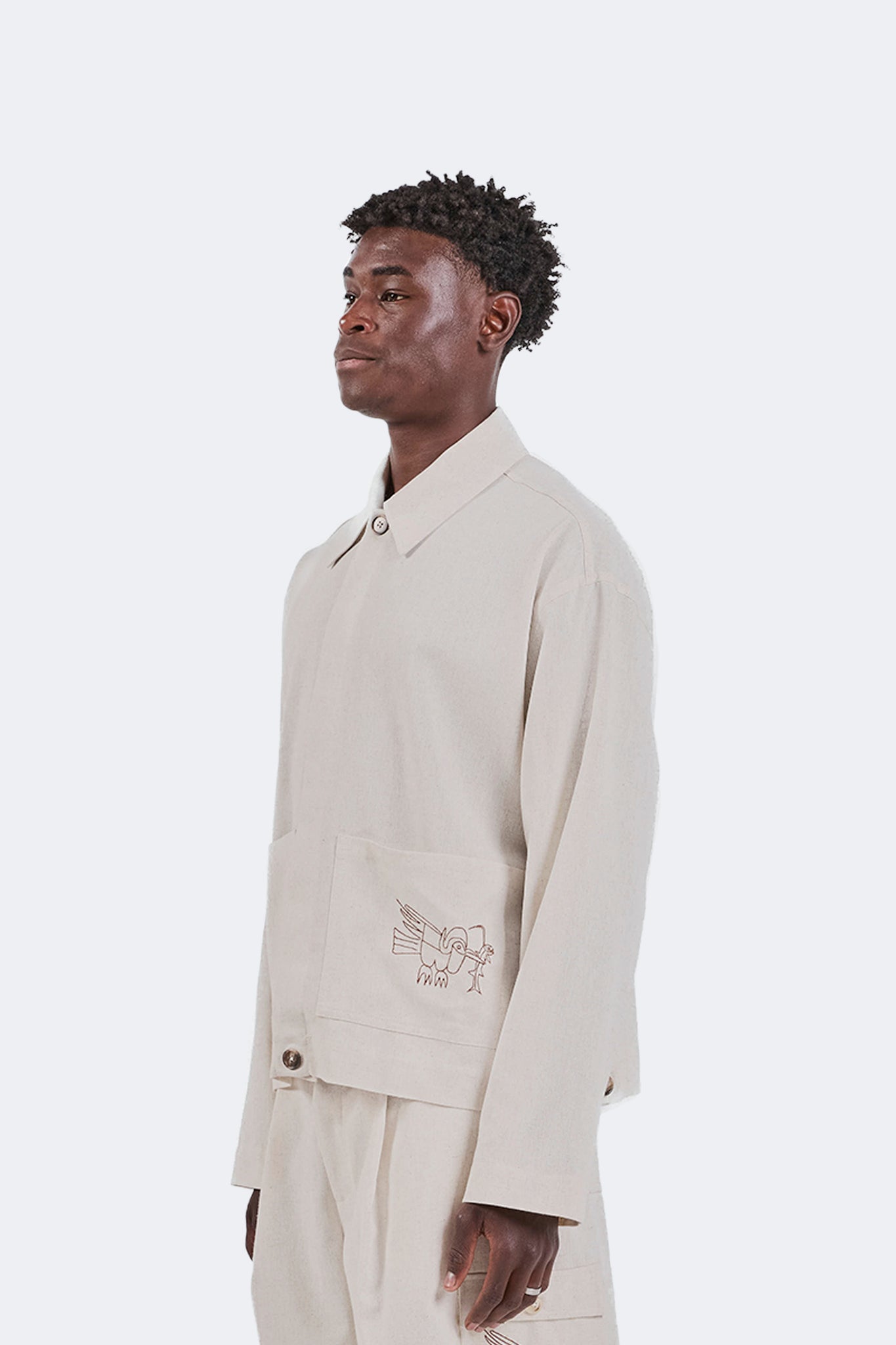 Side detail of the Salango Linen Utility Jacket in Off-White featuring the structured point collar, oversized pocket with embroidered bird motif, and breathable linen construction.
