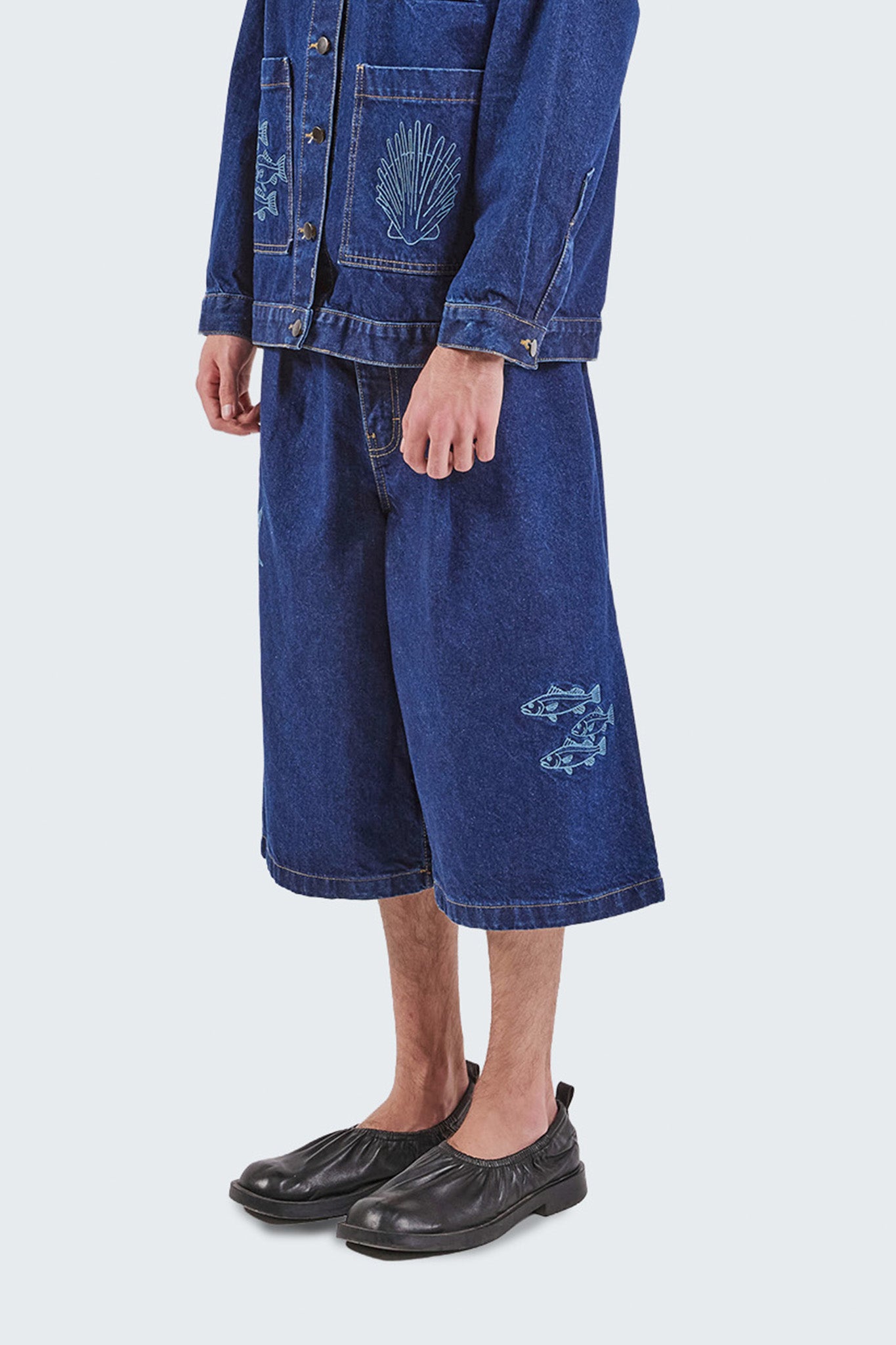 Close-up side detail of the Salango Raw Denim Jorts showcasing embroidered fish motifs, wide-leg construction, raw indigo denim fabric, and cropped length.