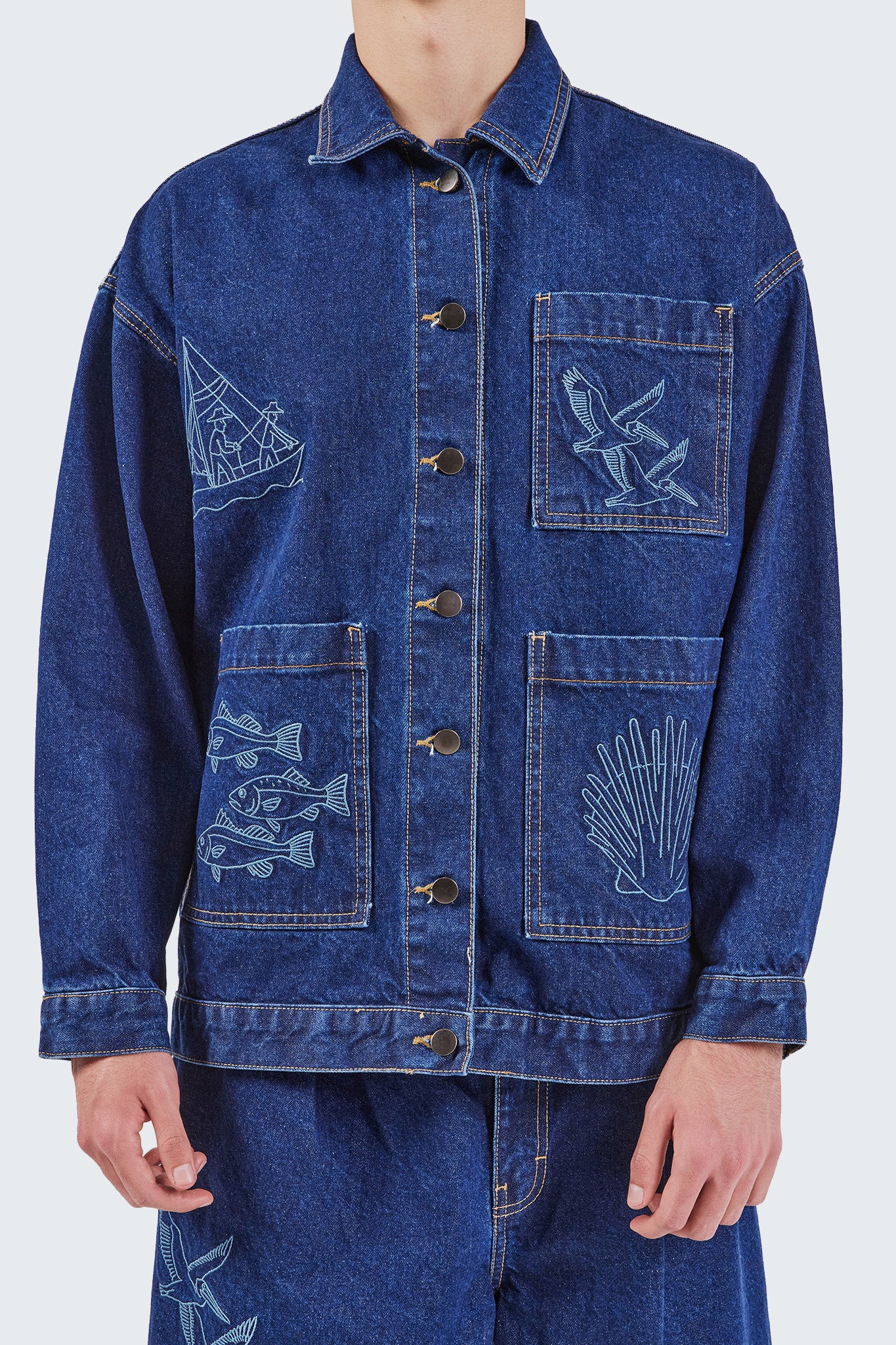 Close-up front view of the Salango Raw Denim Workman Jacket showcasing embroidered coastal motifs including fishermen, seabirds, fish, and shell illustrations, with oversized patch pockets and metal button closure.