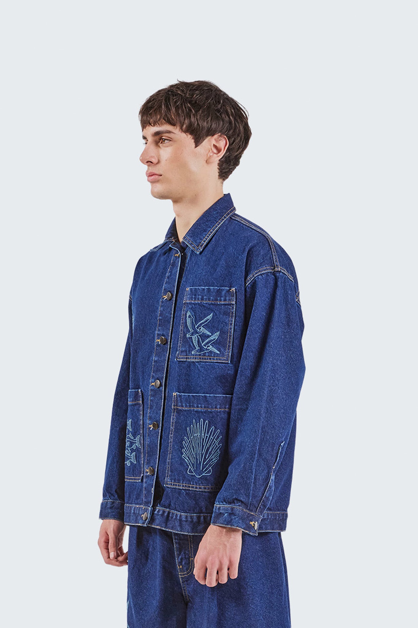 Side view of the Salango Raw Denim Workman Jacket in indigo denim, featuring embroidered seabird and shell motifs, oversized utility pockets, contrast topstitching, and a relaxed workwear-inspired silhouette.