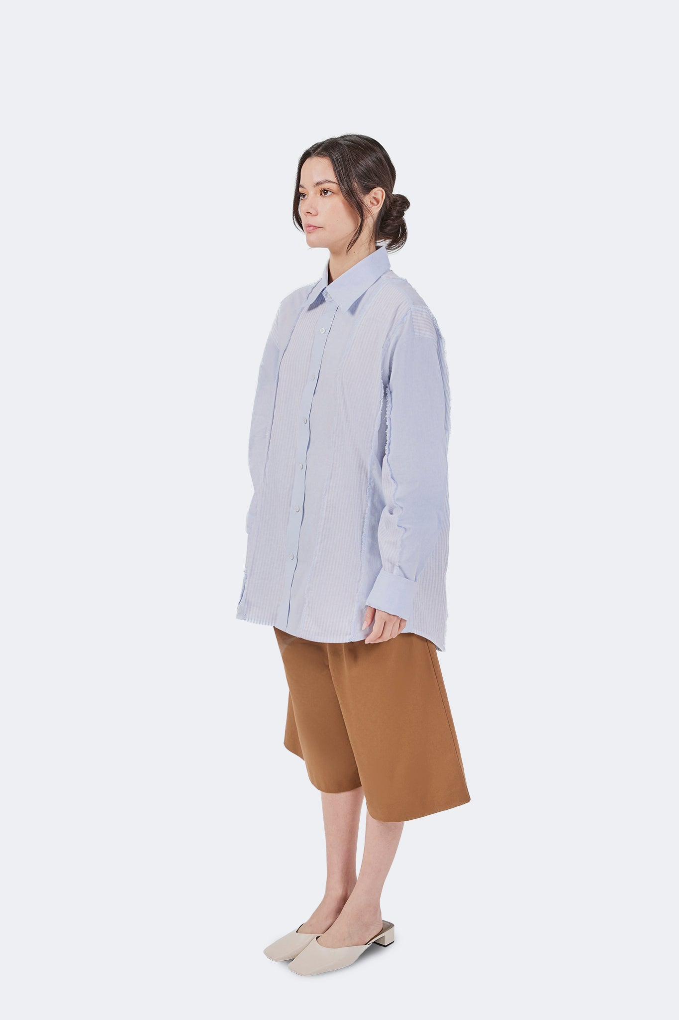 Side view of the Salango Striped Shirt in Sky styled with wide-leg camel shorts, showcasing the relaxed fit, curved hem, and deconstructed panel detailing.