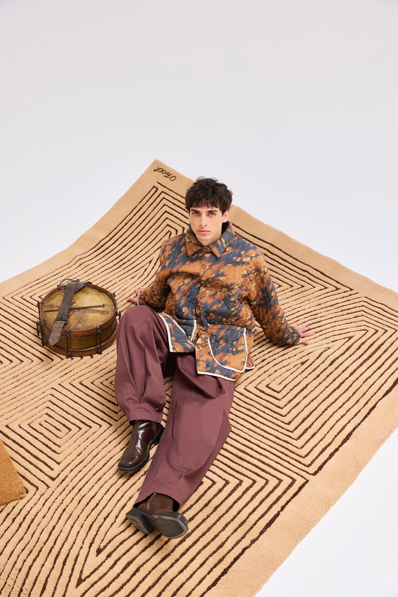 Model wearing the Same Quilted Jacket styled with wide-leg trousers while seated on an Olga Fisch handcrafted rug beside a traditional drum.